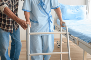Elderly patient with a walker receiving assistance from a nurse in a hospital room.