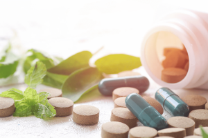 Various tablets and green capsules are scattered around an overturned white bottle, surrounded by fresh green leaves on a white surface.