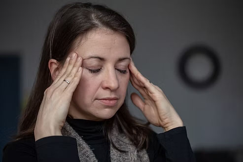 Person in a dark shirt covering their eyes with hands, standing indoors.