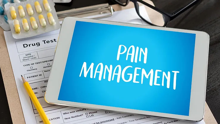The words “pain management” are displayed in a white font in front of a blue background on an iPad screen, next to a tan clipboard, a yellow pencil, and blue and yellow pills.