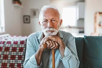 An elderly person sitting, resting hands on a cane, with a cozy home interior in the background.